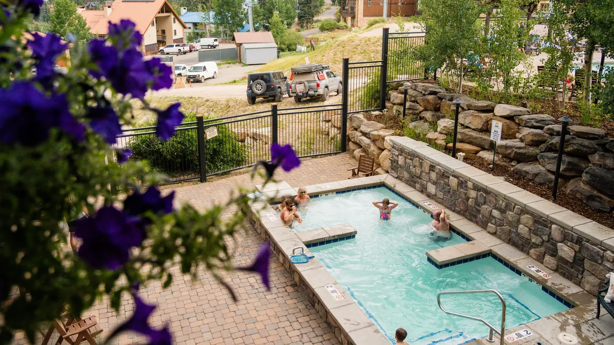 A welcoming hot tub surrounded by vibrant flowers and stone walls, with people unwinding and socializing, set against an outdoor backdrop. The image evokes relaxation after a day of adventure.