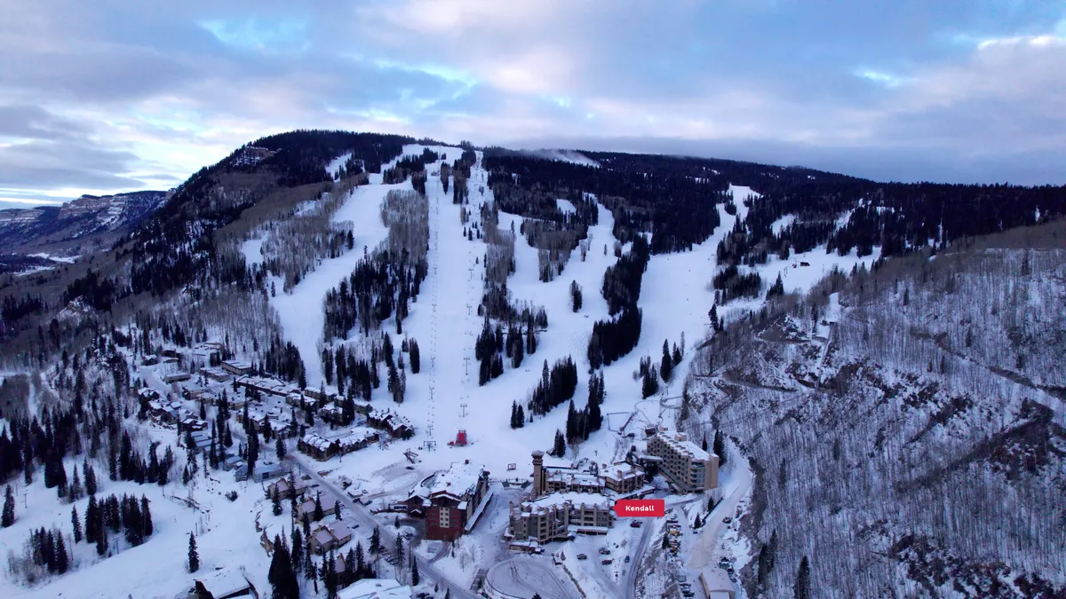 A panoramic view of the resort's snow-covered ski slopes, surrounded by trees and with the warm glow of sunrise or night lighting. Resort buildings and ski lifts are visible, capturing the vibrancy and allure of the area, while suggesting both daytime excitement and relaxing evenings.