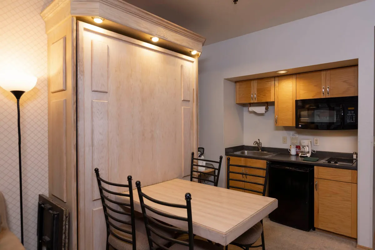 This image showcases a multifunctional interior space featuring a folding table set for dining, adjacent to a compact, well-equipped kitchenette. The scene suggests a cozy atmosphere ideal for preparing and enjoying meals, with the Murphy bed visible in the background to emphasize efficient use of space.