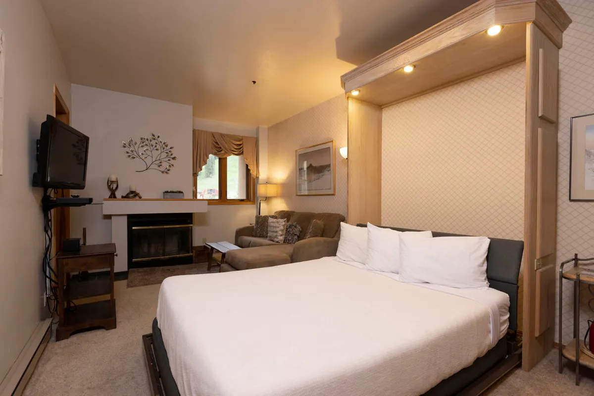 This interior image showcases a cozy bedroom with a plush bed as the centerpiece, accompanied by a comfortable sofa, a warm fireplace, a television, and a window letting in natural light. The overall atmosphere is inviting and emphasizes comfort and relaxation, with soft textures and ambient lighting.