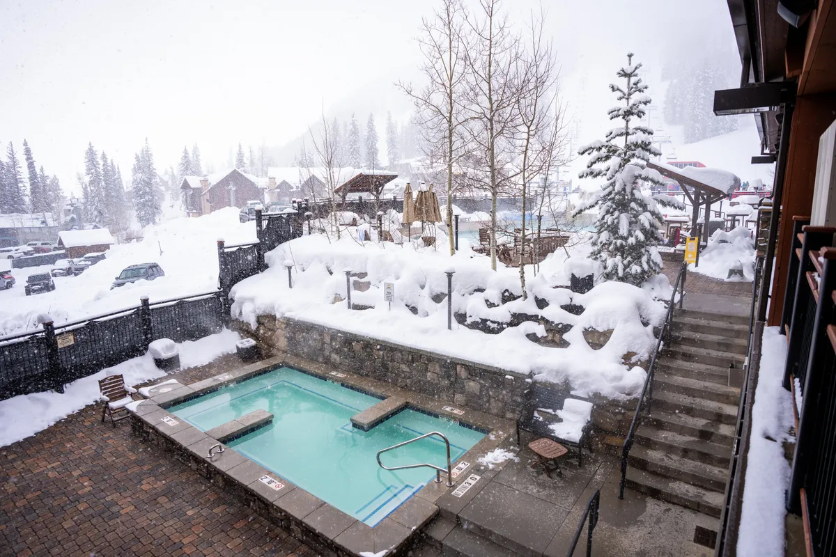 An inviting outdoor hot tub nestled within a snowy landscape, surrounded by trees and nearby resort buildings. Steam rises as people enjoy soaking in the warmth while snowflakes fall, creating an idyllic winter retreat.