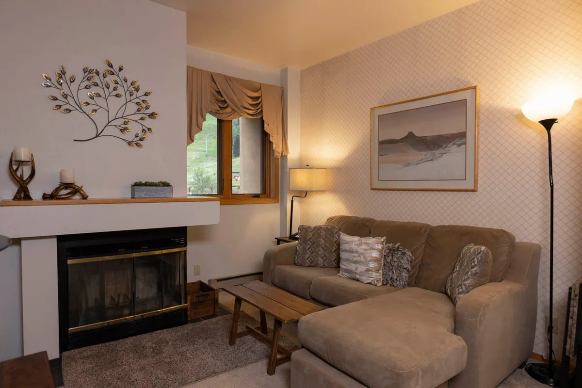 The image features a cozy Durango condo interior, centered around a glowing wood-burning fireplace. A plush sofa invites relaxation, with a soft lamp casting warm light and mountain-inspired wall art enhancing the inviting atmosphere. The nearby window hints at scenic views, while the arrangement suggests an ideal spot for unwinding after a day of outdoor adventure.