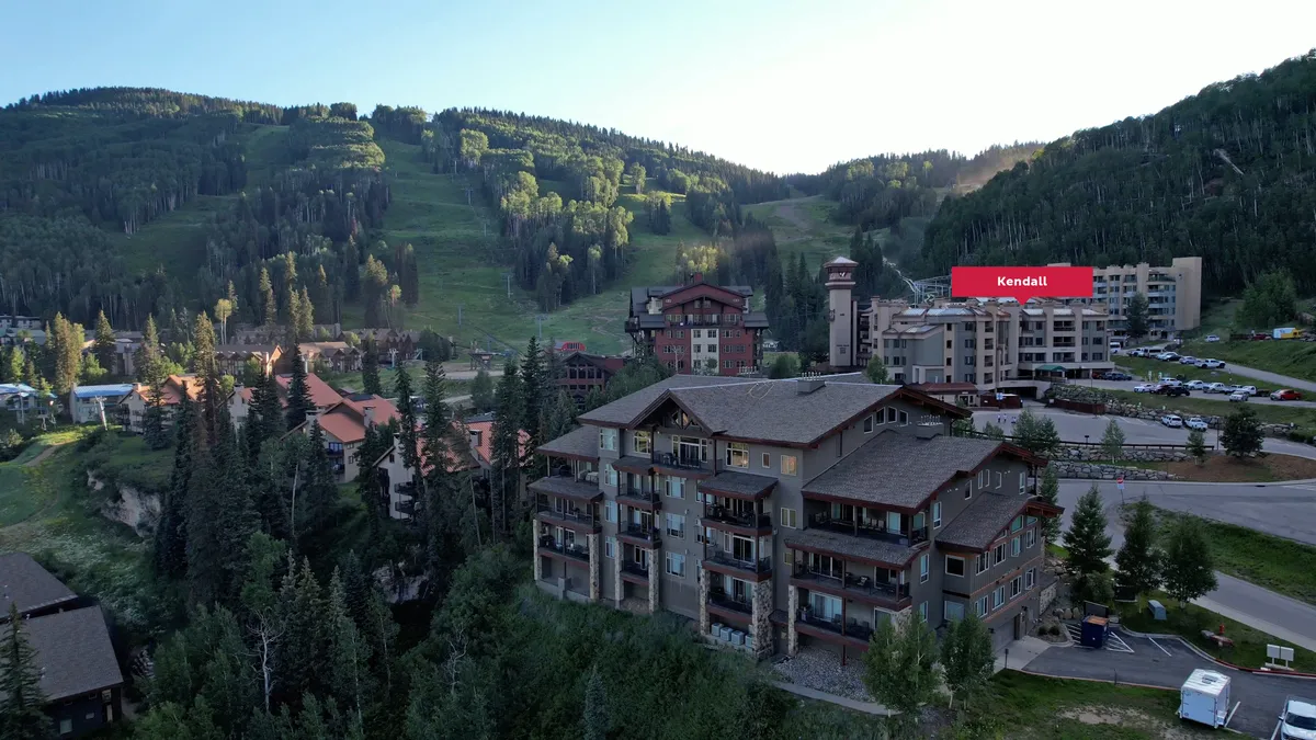 This exterior image features resort buildings nestled against an impressive mountain backdrop, with visible amenities that suggest proximity to outdoor adventures. The setting evokes the excitement of the local area and hints at the activities available nearby, such as skiing, hiking, and mountain biking.