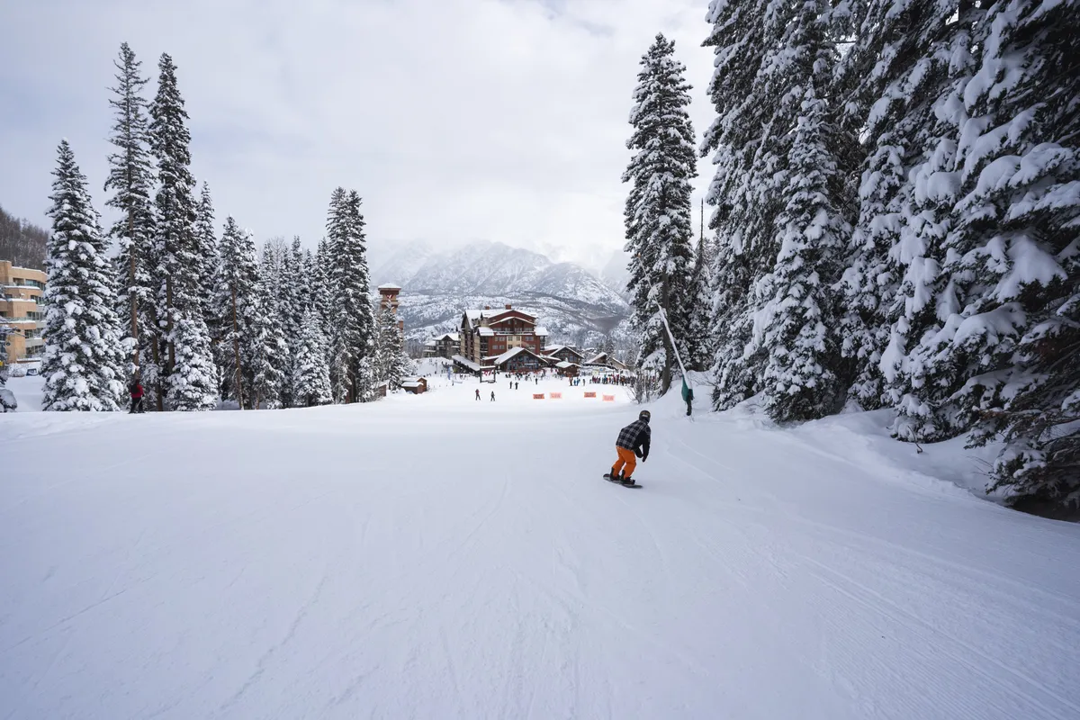A lively exterior scene featuring a snowboarder gliding down a snowy slope, surrounded by snow-covered trees and majestic mountain views. In the background, ski resort buildings nestle at the base of the mountains, while families and friends enjoy outdoor winter activities.