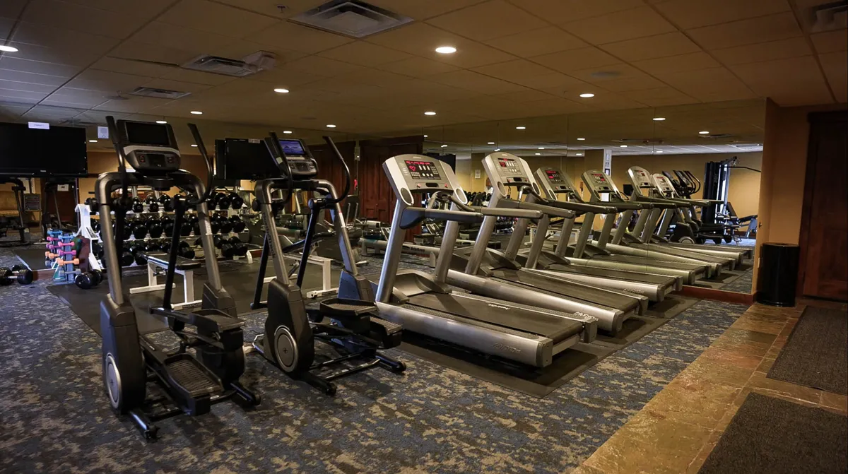 A well-equipped fitness center filled with elliptical machines, treadmills, dumbbells, and mirrors. The space is bright and motivational, with people engaged in workouts and post-exercise socializing.