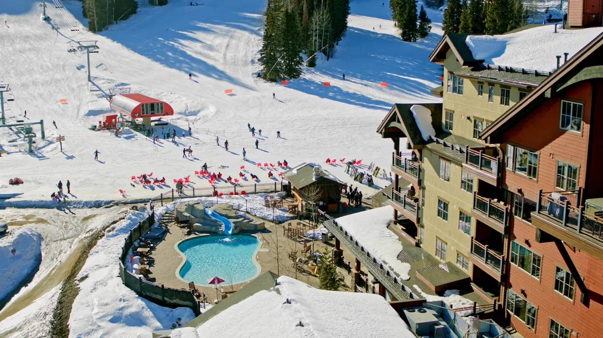 A vibrant exterior scene featuring the lodge's heated swimming pool adjacent to snow-dusted ski slopes, with people skiing, lounging by the pool, and the striking lodge architecture in view. The image captures both recreation and relaxation in a single frame.